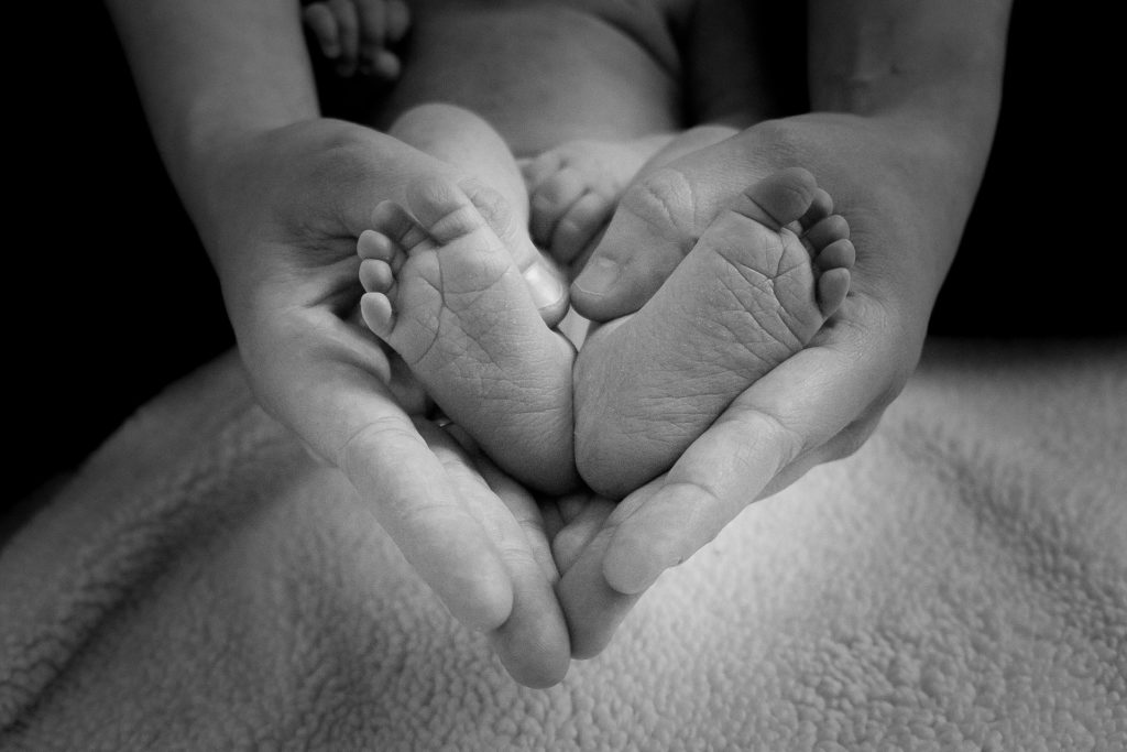 black and white close-up of adult hands cradling the feet of an infant. The hands create the shape of a heart.