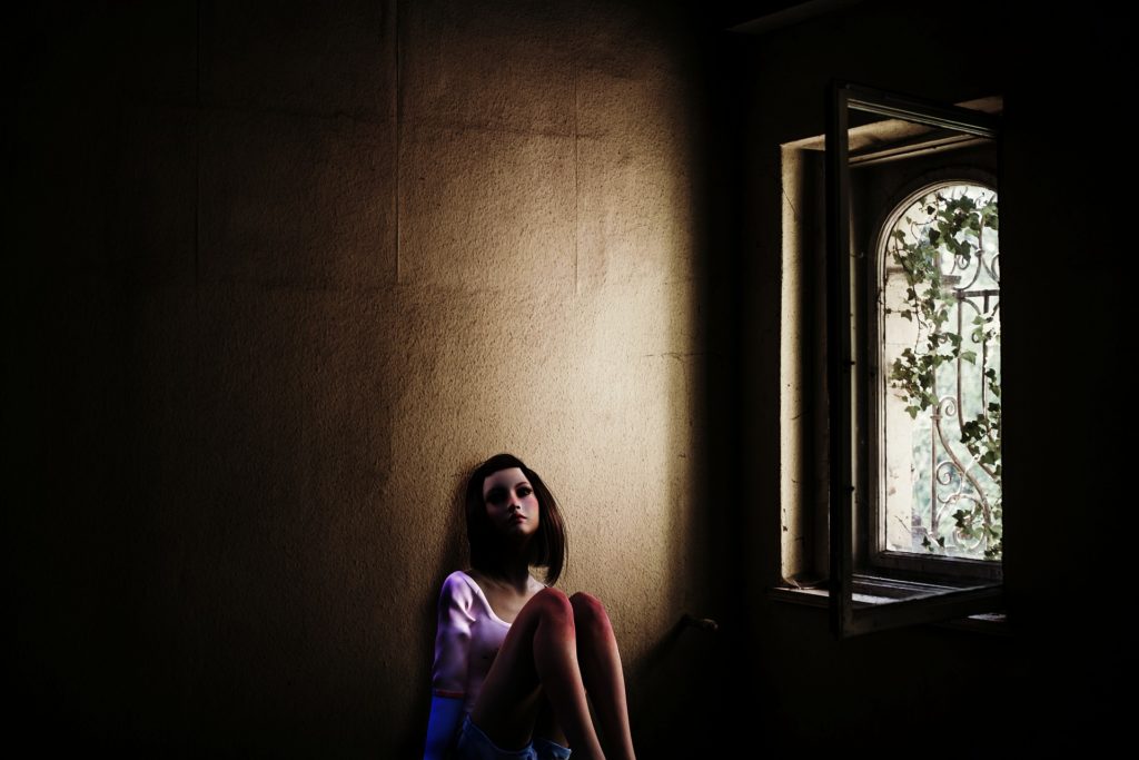 A small beam of light from an arched window shines into a darkened room and on a dark-haired teen girl who leans against the wall with her knees drawn up to her chest.